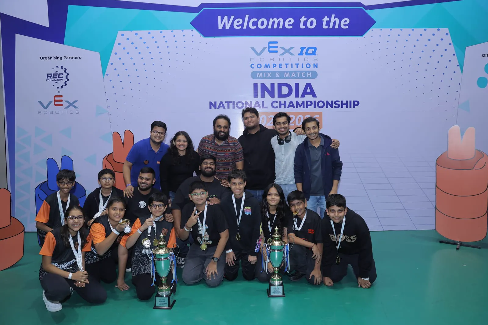 A GSL team and their coaches with two trophies and medals on stage at the VEX IQ Mix and Match India National Championship